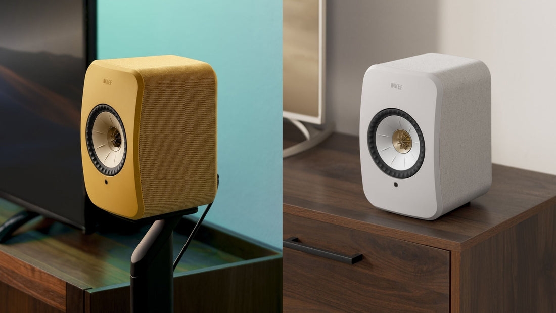 Two KEF LSX II speakers, one yellow and one white, on a wooden surface with a blurred background.