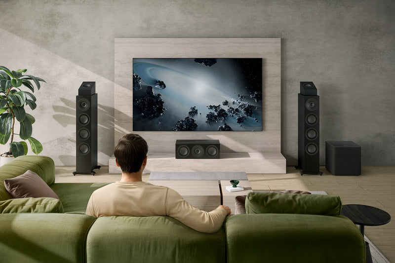 A man sits on a couch watching TV, with KEF Q11 Meta floorstanding speakers, Q8 Meta surround speakers, and a Kube 12 MIE subwoofer nearby.